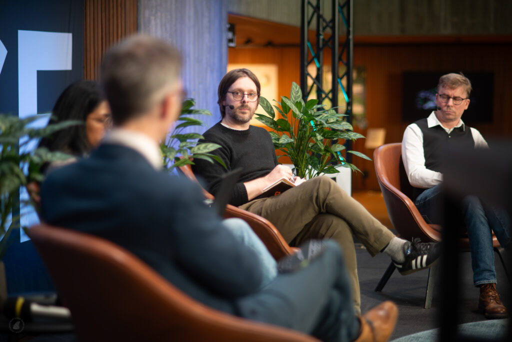 Panelisti Shane Little kuuntelee muistikirja sylissään, ja muita keskustelijoita näkyy etualalla ja vieressä.